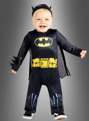 Ein Kleinkind strahlt vor Freude, während es das Original-Batman-Babykostüm trägt – bestehend aus einem schwarzen Anzug mit gelbem Logo, einem gelben Gürtelaufdruck und einer Kopfbedeckung mit Fledermausohren –, und steht vor einem grauen Hintergrund.
