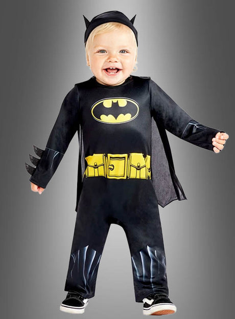 Ein Kleinkind strahlt vor Freude, während es das Original-Batman-Babykostüm trägt – bestehend aus einem schwarzen Anzug mit gelbem Logo, einem gelben Gürtelaufdruck und einer Kopfbedeckung mit Fledermausohren –, und steht vor einem grauen Hintergrund.
