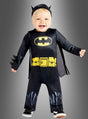 Ein Kleinkind strahlt vor Freude, während es das Original-Batman-Babykostüm trägt – bestehend aus einem schwarzen Anzug mit gelbem Logo, einem gelben Gürtelaufdruck und einer Kopfbedeckung mit Fledermausohren –, und steht vor einem grauen Hintergrund.