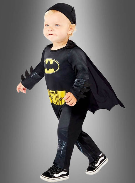 Ein kleines Kind steht vor einem grauen Hintergrund, bereit, in dem entzückenden Original-Batman-Babykostüm die Welt zu retten – ein schwarzer Anzug mit Umhang, gelbem Aufdruck eines Werkzeuggürtels und passenden schwarzen Schuhen, perfekt als Superhelden-Outfit für Kinder.