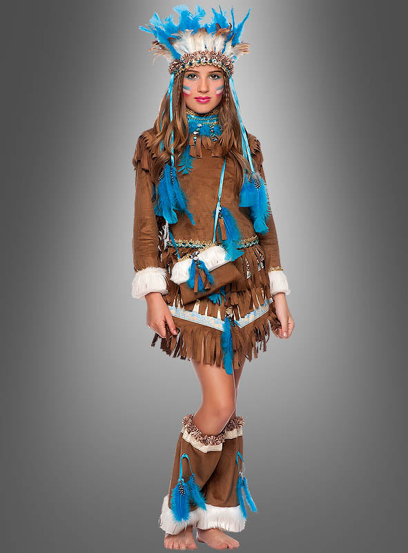 Ein Mädchen trägt das „Indianer-Kleid für Mädchen Deluxe“ aus braunem, mit Fransen verziertem Stoff mit blauen Federakzenten, dazu einen passenden Kopfschmuck und passende Beinhüllen. Sie posiert vor einem grauen Hintergrund, wodurch die farbenfrohen Details dieses bezaubernden Fasching-Kostüms besonders gut zur Geltung kommen.