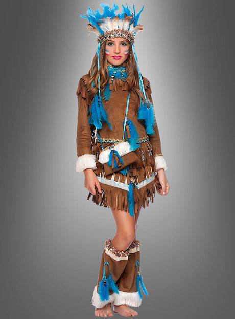 Ein Mädchen trägt das „Indianer-Kleid für Mädchen Deluxe“ aus braunem, mit Fransen verziertem Stoff mit blauen Federakzenten, dazu einen passenden Kopfschmuck und passende Beinhüllen. Sie posiert vor einem grauen Hintergrund, wodurch die farbenfrohen Details dieses bezaubernden Fasching-Kostüms besonders gut zur Geltung kommen.