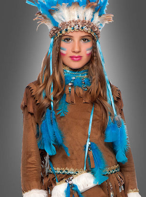 Ein Mädchen, gekleidet im „Indianer-Kleid für Mädchen Deluxe“ – einem braunen, mit Fransen verzierten Outfit, das mit blauen Federn und einem Federkopfschmuck geschmückt ist, die Wangen zur Wirkung bemalt –, steht vor einem grauen Hintergrund und ist in ihrem traditionellen Indianer-Kostüm bereit für den Fasching.