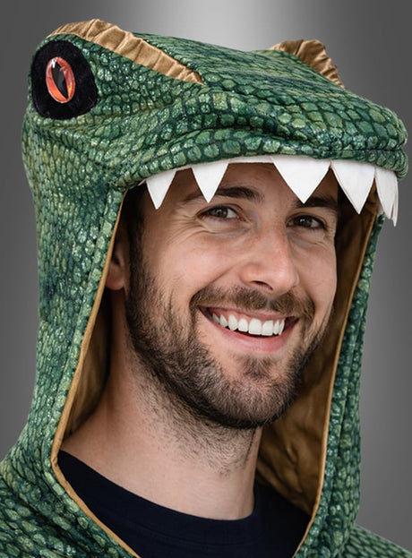 Ein Mann lächelt, während er das T-Rex-Overall-Kostüm für Erwachsene trägt, das sich durch eine grüne Dinosaurierkapuze mit scharfen weißen Zähnen und roten Augendetails vor grauem Hintergrund auszeichnet – perfekt für alle, die auf der Suche nach einem lustigen T-Rex- oder Dino-Kostüm für Erwachsene sind.