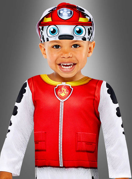Ein kleines Kind lächelt in seinem „Paw Patrol“-Marshall-Kostüm als Dalmatiner, das aus einer roten Weste, weißen Ärmeln mit schwarzen Dalmatinerflecken und einer passenden Mütze besteht, vor einem grauen Hintergrund mit Farbverlauf.