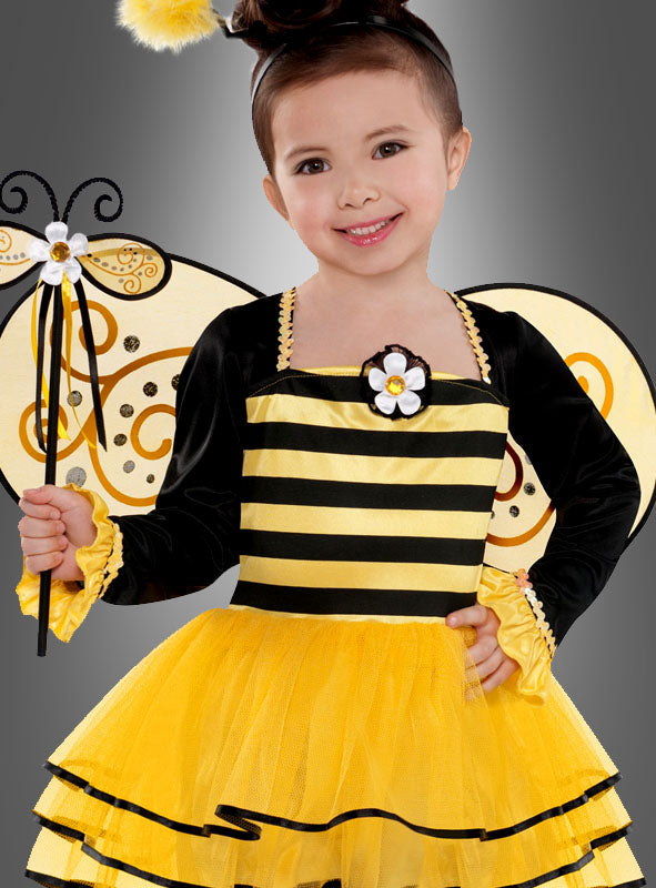 Ein kleines Kind in einem gelb-schwarz gestreiften Bienenkostüm, komplett mit Flügeln und einem Zauberstab, lächelt vor einem grauen Hintergrund in die Kamera – dieses bezaubernde „Fleißiges Bienchen“-Kinderkostüm eignet sich ideal als Faschingskostüm für Kinder oder als Karnevalskostüm für Mädchen.