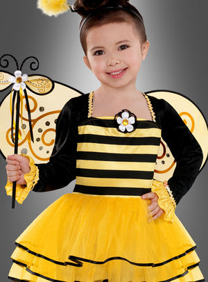 Ein kleines Kind in einem gelb-schwarz gestreiften Bienenkostüm, komplett mit Flügeln und einem Zauberstab, lächelt vor einem grauen Hintergrund in die Kamera – dieses bezaubernde „Fleißiges Bienchen“-Kinderkostüm eignet sich ideal als Faschingskostüm für Kinder oder als Karnevalskostüm für Mädchen.
