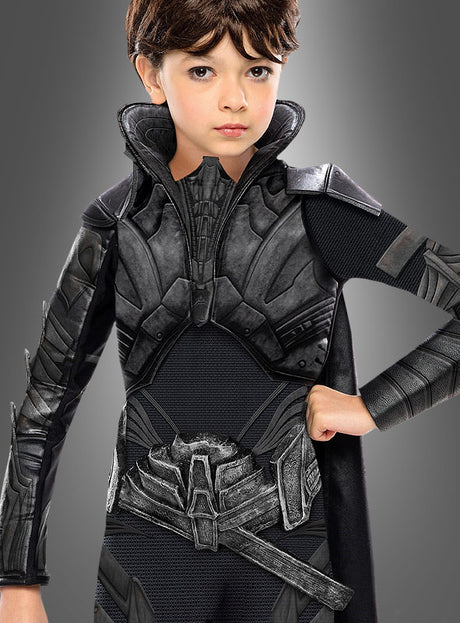 Ein kleines Kind, das das „Faora Man of Steel“-Kinderkostüm trägt – ein detailreiches, schwarzes Rüstungskostüm mit hohem Kragen und Umhang –, steht vor einem grauen Hintergrund.