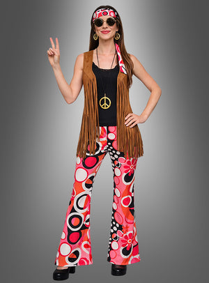 Eine Frau, die das 7-teilige Hippie-Kostüm „Dream Circles“ trägt – bestehend aus einer Weste mit Fransen, einer bunten Schlaghose, einem Stirnband, einer großen Halskette mit Peace-Zeichen und einer runden Sonnenbrille –, zeigt das Peace-Zeichen.