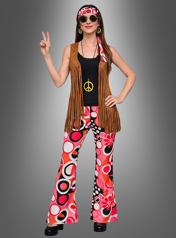 Eine Frau, die das 7-teilige Hippie-Kostüm „Dream Circles“ trägt – bestehend aus einer Weste mit Fransen, einer bunten Schlaghose, einem Stirnband, einer großen Halskette mit Peace-Zeichen und einer runden Sonnenbrille –, zeigt das Peace-Zeichen.