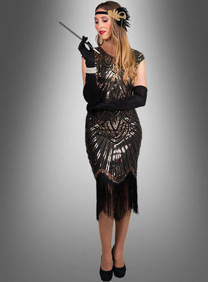 Eine Person, gekleidet im „Funkelndes Gatsby Kleid Daisy“ – einem schwarz-goldenen Paillettenkleid im authentischen Stil der 20er Jahre –, komplett mit Fransen, Stirnband und langen Handschuhen, hält eine Zigarettenspitze in der Hand und steht vor einem schlichten grauen Hintergrund.