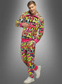 Ein Mann, der den „Cooler Trainingsanzug Neon 80er Flash Herren“ trägt – einen farbenfrohen Trainingsanzug mit bunten, geometrischen Formen in Pink, Gelb und Grün, die an ein kultiges 80er-Jahre-Kostüm erinnern –, steht vor einem grauen Hintergrund mit Farbverlauf und lächelt.