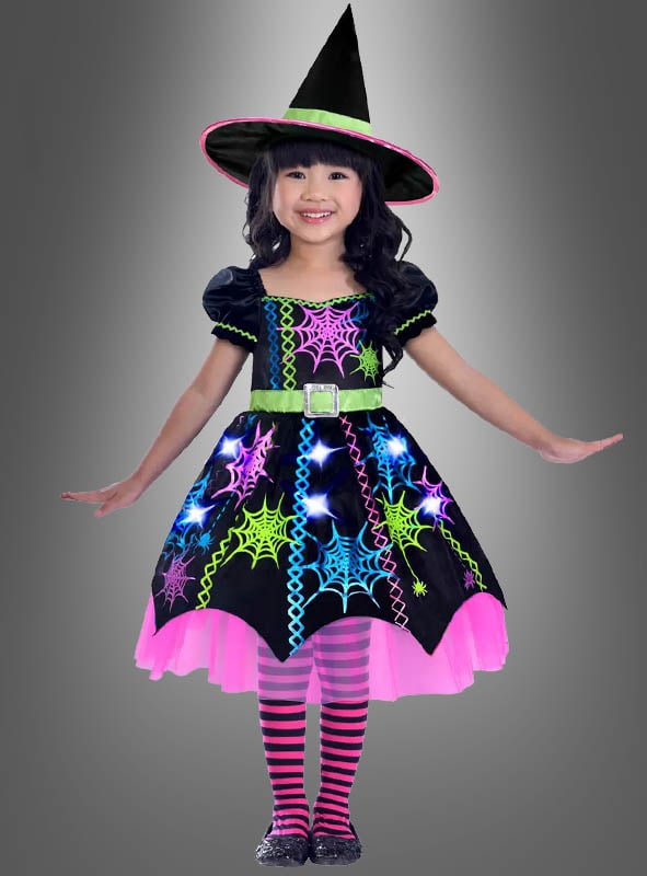 Ein junges Mädchen trägt ein „Bunte Spinnen-Hexe“-Kinderkostüm mit einem spitzen Hut, neonfarbenen Spinnennetzmustern und gestreiften rosa Strumpfhosen und steht vor einem grauen Hintergrund.