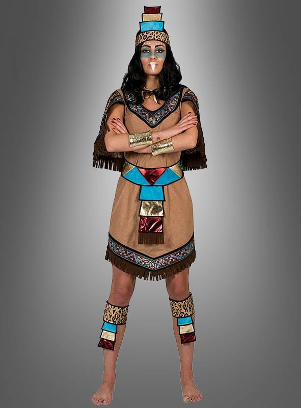 Eine Person, die das „Aztekische Königin“-Kostüm trägt – inspiriert von den Traditionen der amerikanischen Ureinwohner und mit farbenfrohen geometrischen Mustern sowie goldenen Arm- und Beinbändern –, steht barfuß und mit verschränkten Armen vor einem grauen Hintergrund mit Farbverlauf.