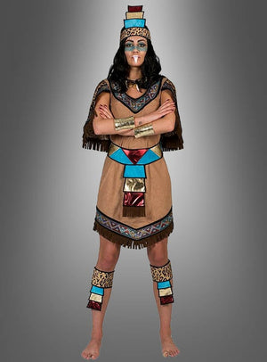 Eine Person, die das „Aztekische Königin“-Kostüm trägt – inspiriert von den Traditionen der amerikanischen Ureinwohner und mit farbenfrohen geometrischen Mustern sowie goldenen Arm- und Beinbändern –, steht barfuß und mit verschränkten Armen vor einem grauen Hintergrund mit Farbverlauf.