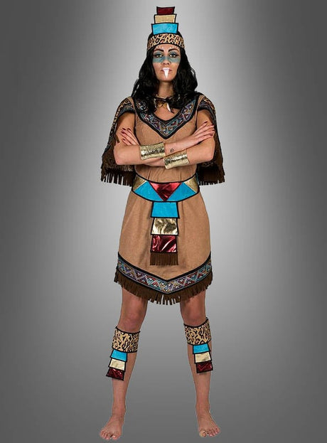 Eine Person, die das „Aztekische Königin“-Kostüm trägt – inspiriert von den Traditionen der amerikanischen Ureinwohner und mit farbenfrohen geometrischen Mustern sowie goldenen Arm- und Beinbändern –, steht barfuß und mit verschränkten Armen vor einem grauen Hintergrund mit Farbverlauf.