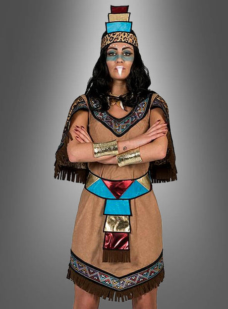 Eine Person, die das „Aztekische Königin“-Kostüm trägt – mit geometrischen Mustern, metallischen Akzenten und blauen, roten und goldenen Formen sowie einem Kopfschmuck und Gesichtsbemalung –, steht mit verschränkten Armen vor einem grauen Hintergrund mit Farbverlauf.