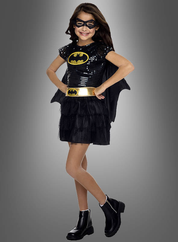 Ein Mädchen im „Batgirl-Kleid für Mädchen“ – einem umwerfenden schwarzen Pailletten-Kinderkostüm mit Maske, Umhang, goldenem Gürtel und schwarzen Stiefeln – steht vor einem grauen Hintergrund; dieses offizielle DC-Comics-Lizenzkostüm ist perfekt für jeden kleinen Superhelden-Fan.
