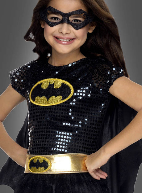 Ein lächelndes Kind trägt das Batgirl-Kleid für Mädchen, ein authentisches Kostüm mit DC-Comics-Lizenz, das mit schwarzen Pailletten, einem goldenen Batman-Logo, einem goldenen Gürtel, einer schwarzen Maske und einem Umhang ausgestattet ist – perfekt für kleine Heldinnen.