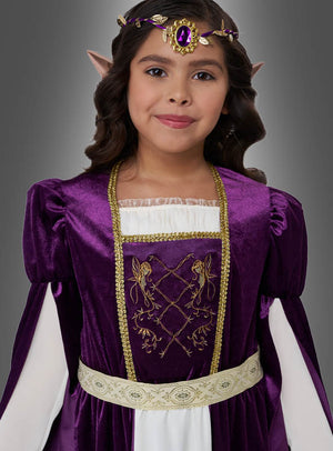 Ein Kind, das das „Elfenkönigin Violetta Kleid lila“ trägt – ein bezauberndes, mittelalterlich anmutendes lila Kleid mit goldenen Verzierungen und einem mit Edelsteinen besetzten Kopfschmuck –, steht vor einem grauen Hintergrund. Spitz zulaufende, elfenhafte Ohren runden dieses zauberhafte „Elfenkönigin Kinderkostüm“-Ensemble ab.