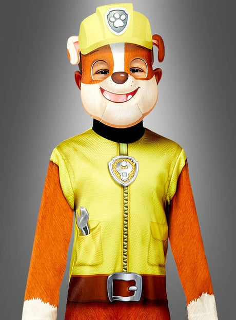 Eine Person, die das Rubble-Kostüm von Paw Patrol trägt – bestehend aus einem gelben Bauhelm, einem Abzeichen und einem braunen Hundekostüm mit offizieller Nickelodeon-Lizenz –, steht vor einem grauen Hintergrund.