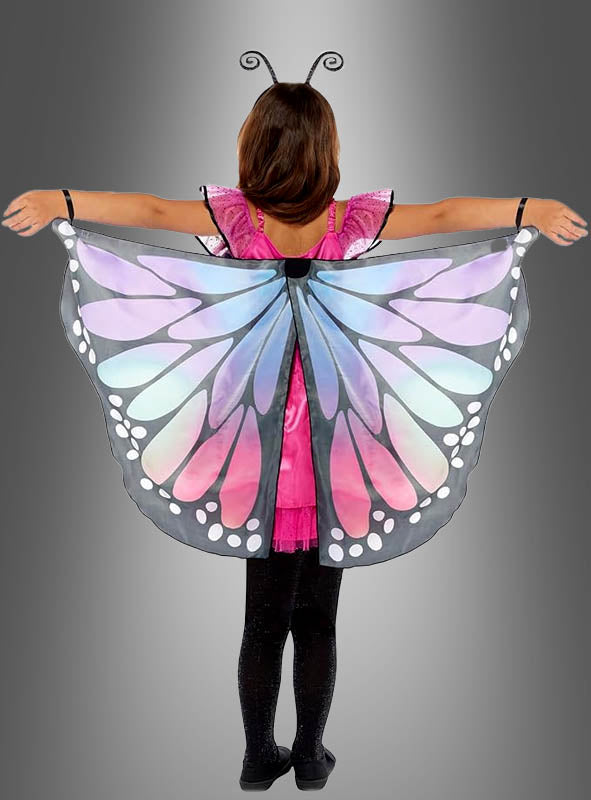 Ein Kind steht mit ausgestreckten Armen vor einem grauen Hintergrund und trägt schwarze Leggings, ein rosa Schmetterlingskleid und farbenfrohe Schmetterlingsflügel mit blauen, violetten und weißen Mustern – ein bezauberndes Schmetterlings-Traumkostüm für Kinder, das sich perfekt für fantasievolles Spielen eignet.