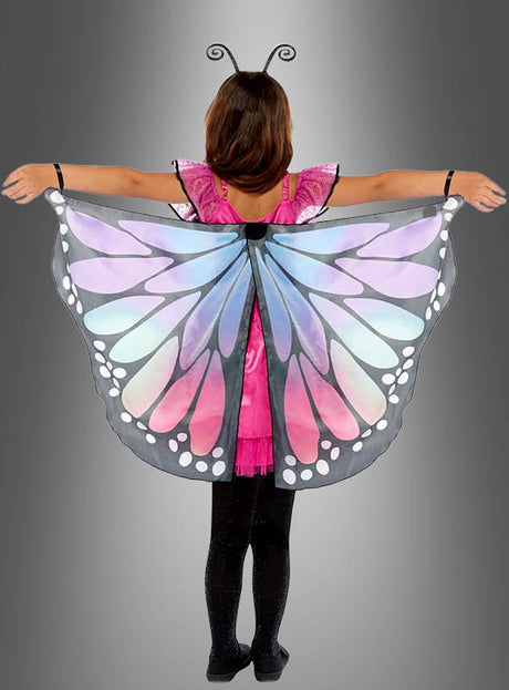 Ein Kind steht mit ausgestreckten Armen vor einem grauen Hintergrund und trägt schwarze Leggings, ein rosa Schmetterlingskleid und farbenfrohe Schmetterlingsflügel mit blauen, violetten und weißen Mustern – ein bezauberndes Schmetterlings-Traumkostüm für Kinder, das sich perfekt für fantasievolles Spielen eignet.