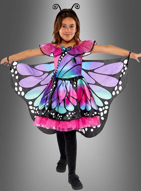 Ein Mädchen im „Schmetterling-Traumkostüm für Kinder“ – bestehend aus einem bunten Kleid mit leuchtend schwarzen, violetten, rosa und blauen Schmetterlingsflügeln, dazu schwarze Leggings, schwarze Schuhe und ein Stirnband mit Fühlern – steht mit ausgestreckten Armen vor einem grauen Hintergrund; perfekt als Kinderkostüm.