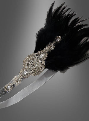 Ein dekoratives Haarband im Stil der 20er Jahre mit Strass und Perlen namens „Lilli“, verziert mit schwarzen Federn und einem kunstvollen Design aus silbernen Perlen, das an eleganten Kopfschmuck der 20er Jahre erinnert, dargestellt vor einem grauen Hintergrund.