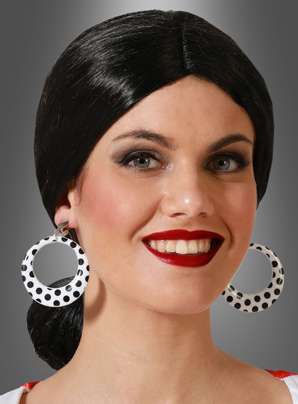 Eine Frau mit dunklem Haar, das zu einem tief sitzenden Dutt zusammengebunden ist, trägt die „Polka Dot Ohrringe 50er Jahre“ – große, weiße Ohrringe mit Polka-Dot-Muster, die vom Stil der 1950er Jahre inspiriert sind – sowie roten Lippenstift und ein weißes Oberteil. Vor einem grauen Hintergrund mit Farbverlauf lächelt sie in die Kamera.