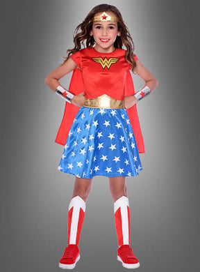 Ein junges Mädchen, das ein klassisches Wonder-Woman-Kinderkostüm trägt – bestehend aus einem roten Umhang, einem blauen Rock mit Sternenmuster, einem goldenen Gürtel und roten Stiefeln –, steht vor einem grauen Hintergrund.