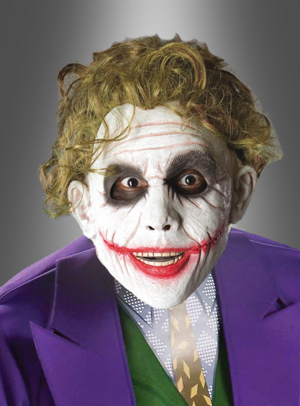 Eine Person mit weißem Gesichts-Make-up, geschwärzten Augen und roten Lippen, die zu einem breiten Grinsen verzogen sind, sowie einer violetten Jacke und einer grünen Weste ähnelt einem Comic-Bösewicht, der in einem auffälligen Joker-Kostüm, komplett mit der Original-Joker-Perücke, zum Leben erweckt wurde.