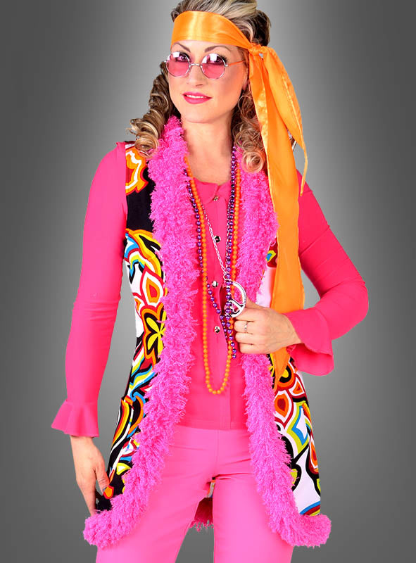 Eine Frau in einem knallpinken Outfit im Stil der 1970er Jahre, bestehend aus der „Hippie Weste Neon Psychodelic“, ergänzt durch ein orangefarbenes Stirnband, eine rosa Sonnenbrille und Perlenketten, steht vor einem grauen Hintergrund – ein kultiges 70er-Jahre-Kostüm, perfekt für den Karneval.