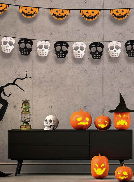 Ein modernes Zimmer, das zu Halloween mit Kürbislaternen, einem Totenkopf, einer Laterne, einem Hexenhut und den festlichen Halloween-Girlanden mit Kürbis- oder Totenkopfmotiven geschmückt ist, die an der Betonwand hängen.