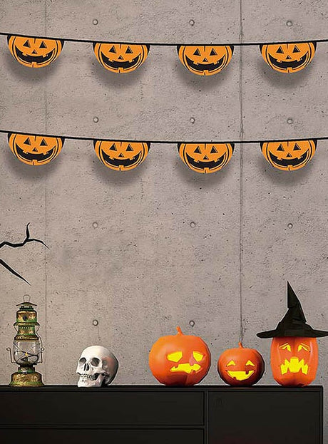 Halloween-Dekoration: Halloween-Girlanden mit Kürbissen oder Totenköpfen, die an einer grauen Wand hängen, drei geschnitzte Kürbisse (einer davon mit einem Hexenhut), eine Laterne, ein künstlicher Totenschädel und ein schwarzer Ast, die auf einem schwarzen Schrank ausgestellt sind.