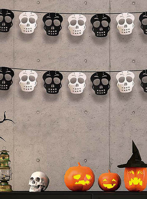 An einer grauen Wand über einem Tisch, auf dem geschnitzte Kürbisse, ein künstlicher Totenkopf, eine Laterne und ein Hexenhut stehen, hängen zwei Reihen schwarz-weißer Halloween-Girlanden mit Kürbis- oder Totenkopfmotiven.