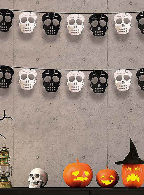 An einer grauen Wand über einem Tisch, auf dem geschnitzte Kürbisse, ein künstlicher Totenkopf, eine Laterne und ein Hexenhut stehen, hängen zwei Reihen schwarz-weißer Halloween-Girlanden mit Kürbis- oder Totenkopfmotiven.