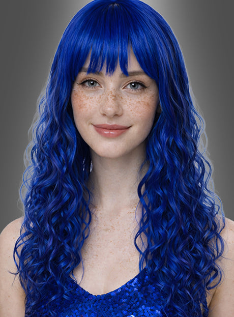 Eine Frau mit langen, lockigen, leuchtend blauen Haaren und Pony – gestylt mit der farbigen Damenperücke „Feline“ von Wellige – trägt ein blaues Pailletten-Oberteil und lächelt leicht vor einem grauen Hintergrund mit Farbverlauf.