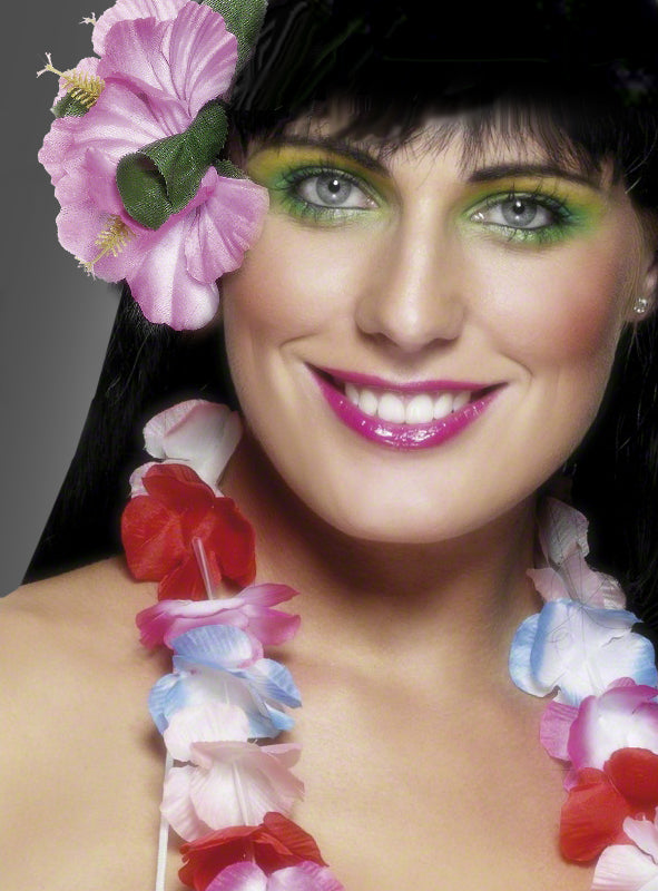 Eine Frau, die „Hawaii Blumen“-Haarschmuck – ein farbenfrohes Haaraccessoire mit Blumenmotiv – trägt, dazu eine rosa Blume im Haar, leuchtend grünen Lidschatten und rosa Lippenstift, lächelt in die Kamera und präsentiert so den perfekten Look für ein Hawaii-Kostüm oder eine Hawaii-Party.