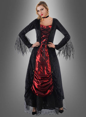 Eine Frau, die das „Vampirin Selina Kleid mit Spitze“ trägt – ein auffälliges schwarz-rotes Kleid im Gothic-Stil mit Spitzenärmeln –, steht mit den Händen in den Hüften vor einem grauen Hintergrund mit Farbverlauf und ist damit das perfekte Halloween-Kostüm als Vampirin oder Halloween-Abendkleid für Damen für eine unvergessliche Nacht.