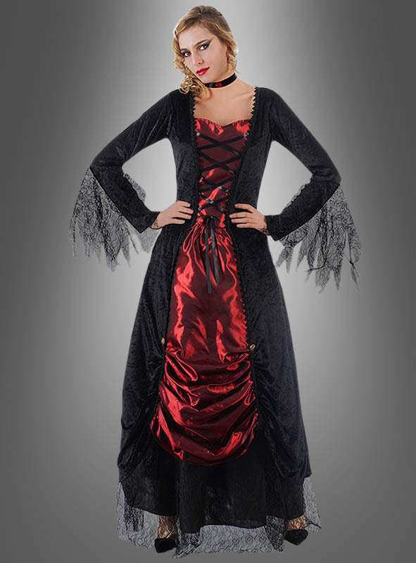 Eine Frau, die das „Vampirin Selina Kleid mit Spitze“ trägt – ein auffälliges schwarz-rotes Kleid im Gothic-Stil mit Spitzenärmeln –, steht mit den Händen in den Hüften vor einem grauen Hintergrund mit Farbverlauf und ist damit das perfekte Halloween-Kostüm als Vampirin oder Halloween-Abendkleid für Damen für eine unvergessliche Nacht.