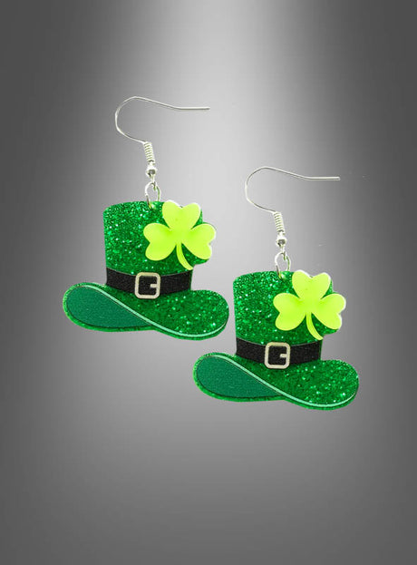 Ein Paar St. Patrick’s Day-Ohrringe in Kleeblattform – grüne, glitzernde Ohrringe in Form von Leprechaun-Hüten mit gelben Kleeblättern und schwarzen Hutbändern – vor einem grauen Hintergrund mit Farbverlauf.
