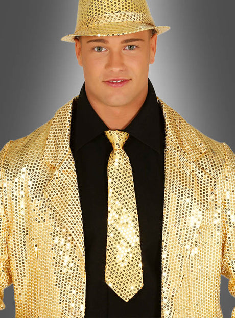 Eine Person steht selbstbewusst vor einem grauen Hintergrund mit Farbverlauf und trägt einen goldenen Paillettenhut sowie eine Jacke mit der schimmernden „Pailletten Krawatten Unisex“ über einem schwarzen Hemd – ein auffälliges Karnevalsaccessoire, das sich perfekt für jede Feier eignet.