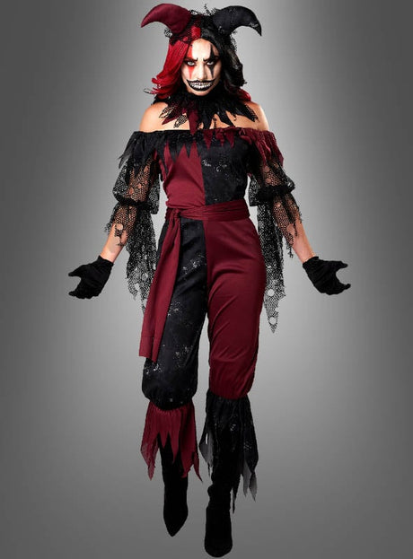 Eine Person, die ein rot-schwarzes Narrenkostüm für Damen mit roten und schwarzen Hörnern, dunklem Make-up und Handschuhen trägt, steht vor einem schlichten grauen Hintergrund.