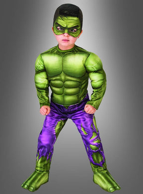 Ein Kind trägt stolz das Hulk-Kostüm für Kleinkinder und präsentiert dabei einen grünen, muskulösen Anzug, lila zerrissene Hosen und grüne Gesichtsbemalung vor einem grauen Hintergrund mit Farbverlauf.