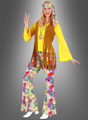 Eine Frau im Hippie-Look, gekleidet im Bettina-Damenkostüm, bestehend aus einem leuchtend gelben Oberteil, einer braunen Weste mit Fransen, einer bunt gemusterten Schlaghose, einem passenden Stirnband und einer Peace-Halskette – perfekt, um den ikonischen Stil der 70er Jahre nachzuahmen –, steht vor einem grauen Hintergrund und zeigt das Peace-Zeichen.