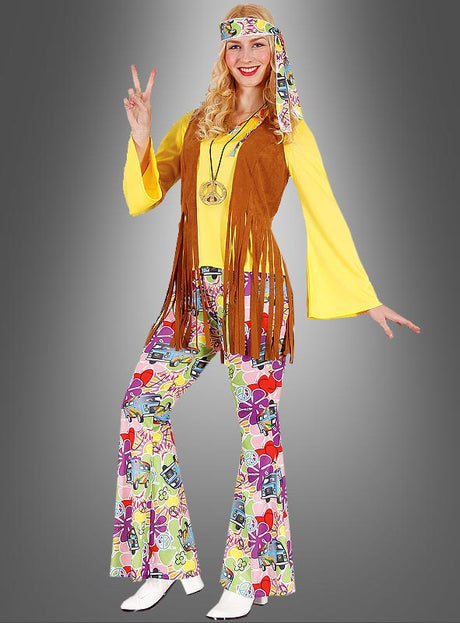 Eine Frau im Hippie-Look-Damenkostüm „Bettina“ – bestehend aus einem gelben Hemd, einer braunen Weste mit Fransen, einer bunten Schlaghose, einem Stirnband und einer Halskette mit Peace-Zeichen – posiert vor einem schlichten grauen Hintergrund. Dieses farbenfrohe Hippie-Damenkostüm verkörpert perfekt den Geist des 70er-Jahre-Kostümstils.