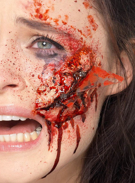 Eine Nahaufnahme des Gesichts einer Person mit schweren, blutigen Verletzungen und einem sichtbaren Glassplitter, der in der Wange steckt. Der realistische „Glassplitter-Unfallwunde“-Make-up-Effekt aus Latex wird durch fachmännisch aufgetragenes Wund-Make-up ergänzt, wodurch der offene Mund und die freiliegenden Zähne erschreckend lebensecht wirken.