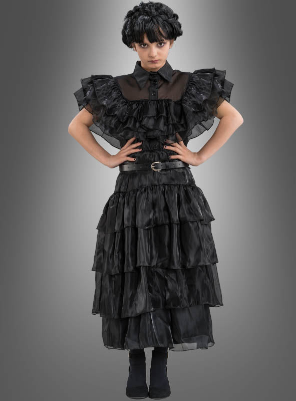 Eine Person steht mit den Händen in den Hüften vor einem grauen Hintergrund mit Farbverlauf und trägt das „Wednesday Mädchen Ballkleid“ – ein mehrlagiges schwarzes Rüschenkleid mit hohem Kragen, breitem Gürtel, dunkler Perücke und schwarzen Schuhen –, perfekt für alle, die auf der Suche nach einem authentischen Wednesday-Kostüm oder einem stilvollen Ballkleid für Mädchen sind, das von ikonischen Figuren inspiriert ist.