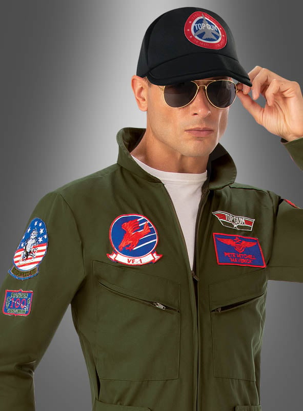 Eine Person, gekleidet in das „Top Gun Pilot mit Namensschild“-Original-Lizenzkostüm – einen grünen Fluganzug mit Aufnähern –, kombiniert mit einer Pilotenbrille und einer schwarzen Mütze mit dem Top-Gun-Logo, steht vor einem grauen Hintergrund mit Farbverlauf.